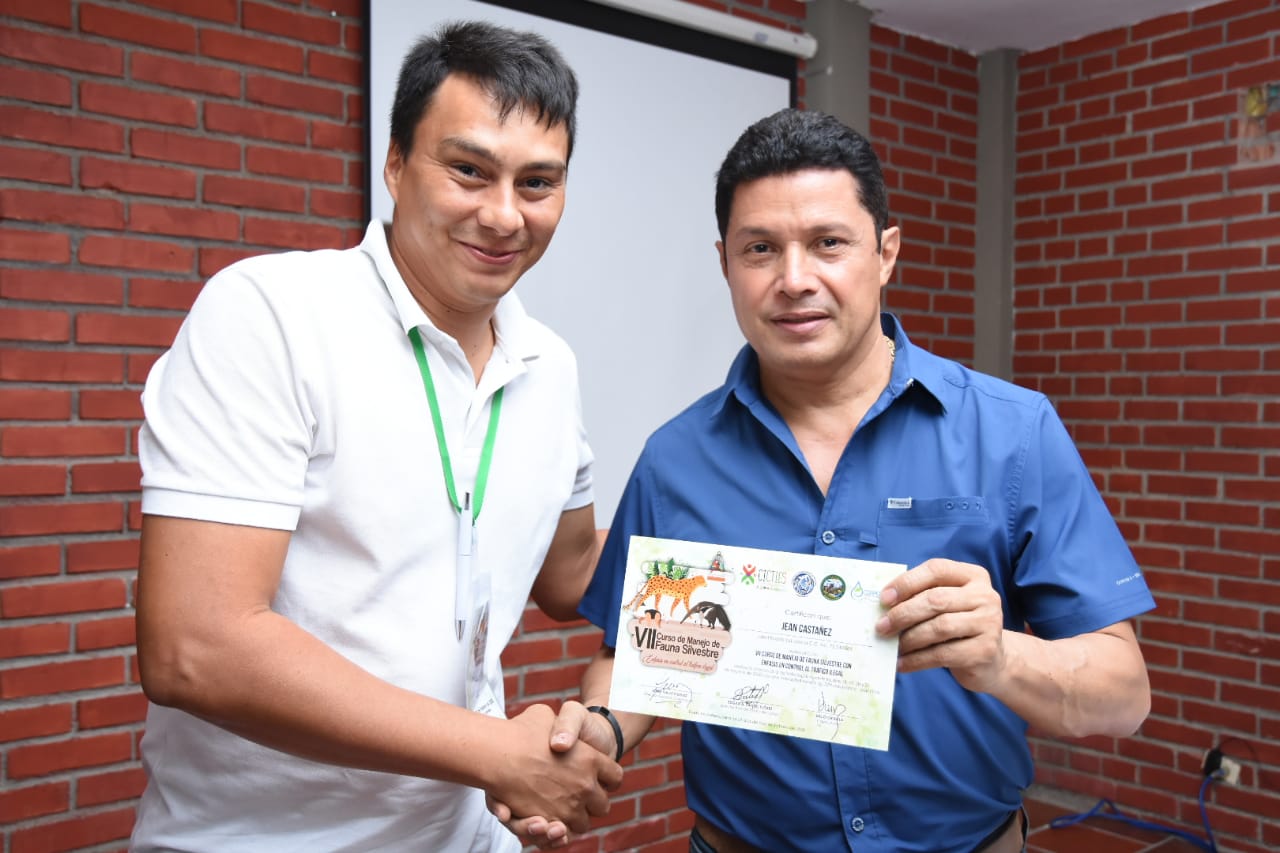 Un hombre entrega un certificado a un participante del curso, dándose la mano, frente a una pared de ladrillo