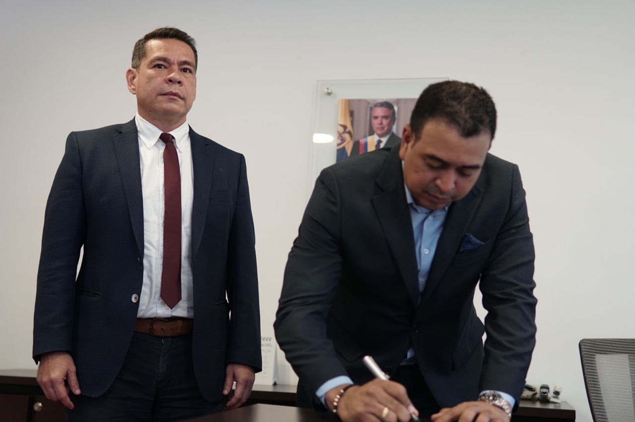 Dos hombres con traje en un despacho, uno de ellos firma un documento durante la posesión del director
