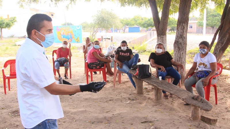 Un funcionario con guantes habla ante los habitantes sentados bajo los árboles durante la capacitación