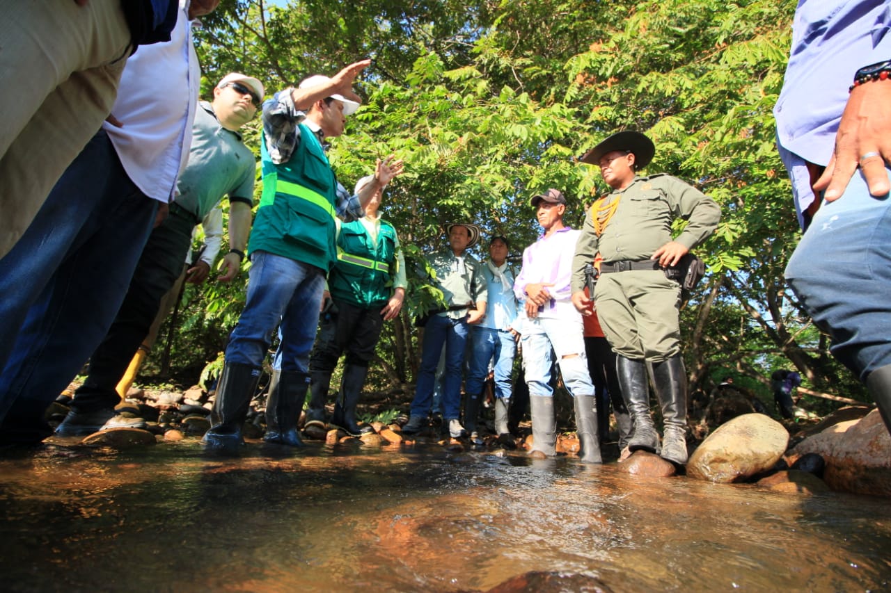 El director y funcionarios con chaleco verde recorren una fuente hídrica con los pies en el agua