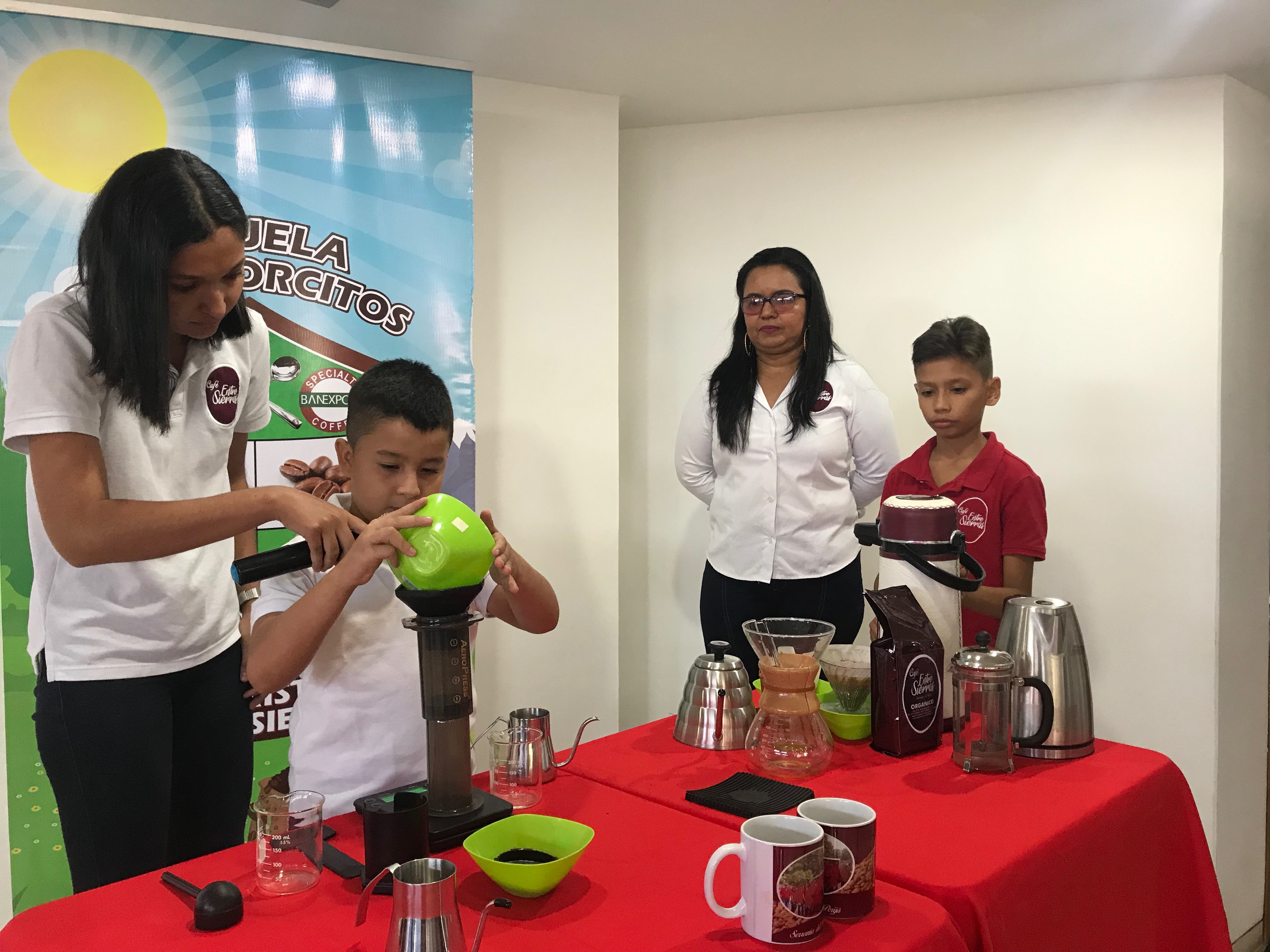 Una joven y un niño preparan café con equipos de filtrado sobre una mesa roja, frente a un pendón de la «Escuela Catadorcitos».