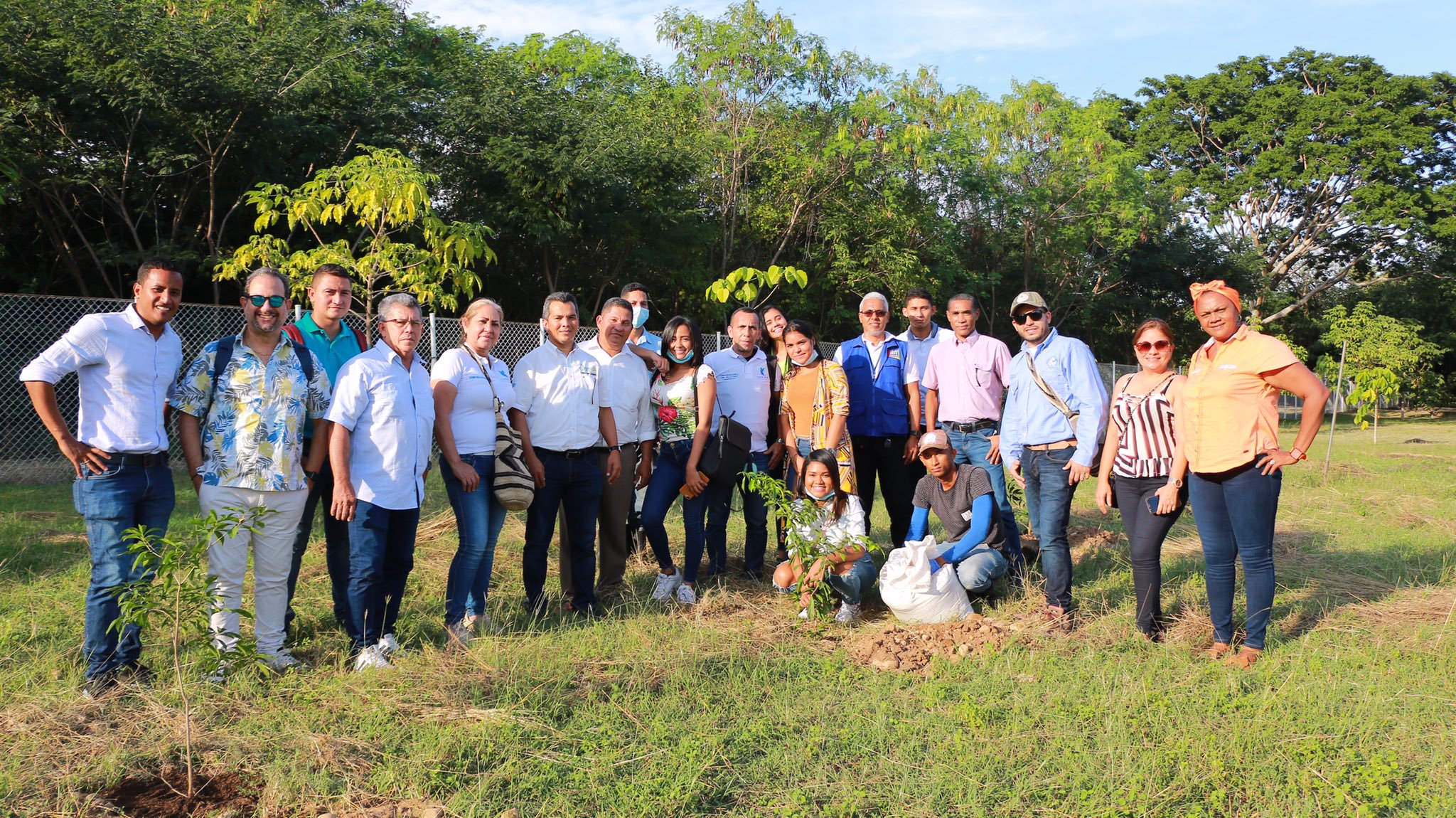 Grupo de personas posa de pie en un terreno con árboles; al frente, una joven agachada junto a un arbolito recién sembrado