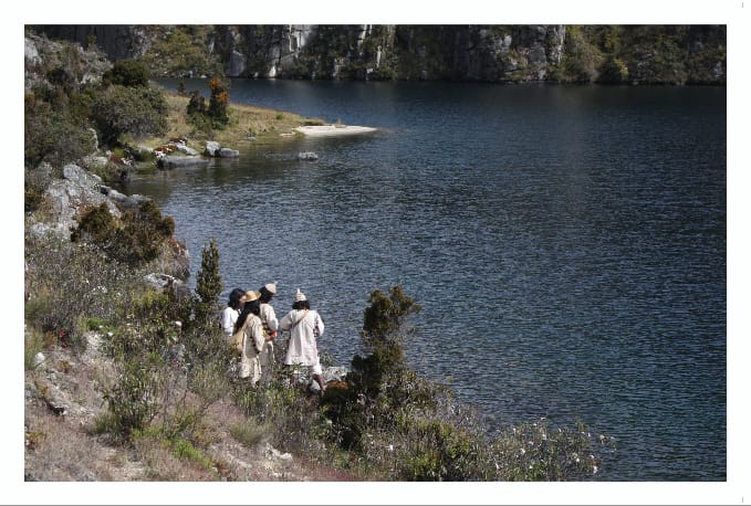 Tres personas con ropa blanca tradicional y sombreros cónicos junto a una laguna de montaña.