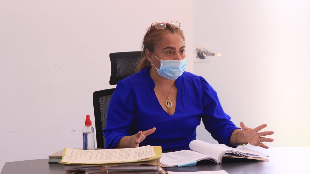 Una funcionaria con blusa azul habla en una oficina junto a documentos sobre el escritorio
