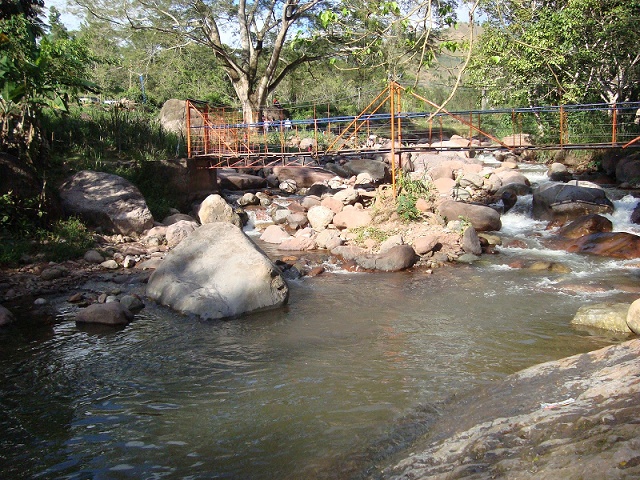 Fotografía del río Manaure: corriente de agua entre grandes rocas, con un puente colgante y árboles al fondo.