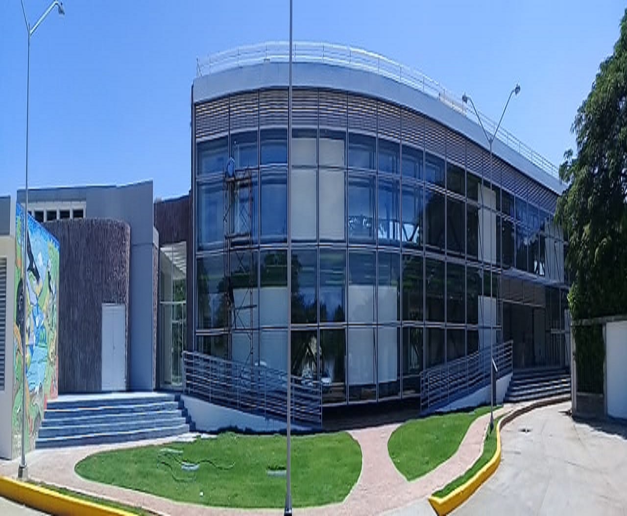 Fachada curva de vidrio y estructura metálica de la sede bioclimática de Corpocesar, con escalinatas, rampas y un mural a la izquierda
