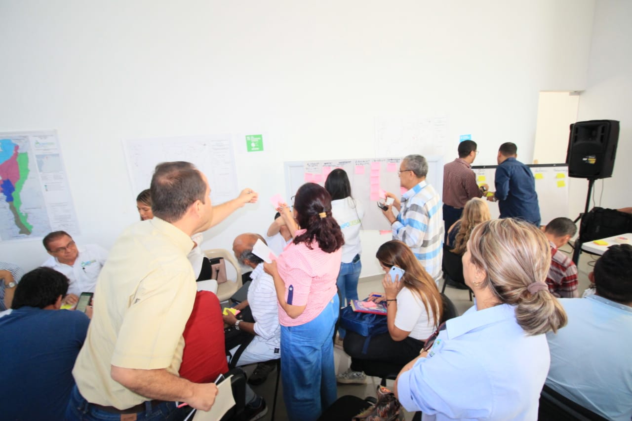 Asistentes a la mesa de trabajo con la comunidad sentados en un salón con mapas en la pared